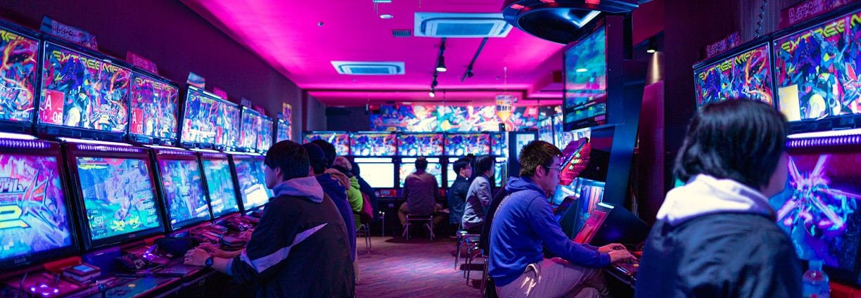 A vibrant arcade with rows of gaming machines adorned with colorful screens. Several people are immersed in playing games, seated on stools in a dimly lit room accented by an intense pink ceiling light. The atmosphere is lively and engaging.