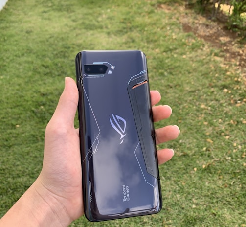 A hand holding a sleek, dark-colored smartphone with geometric patterns and the text 'Tencent Games' on the back cover. The background features a grassy area with some trees or bushes on the side.